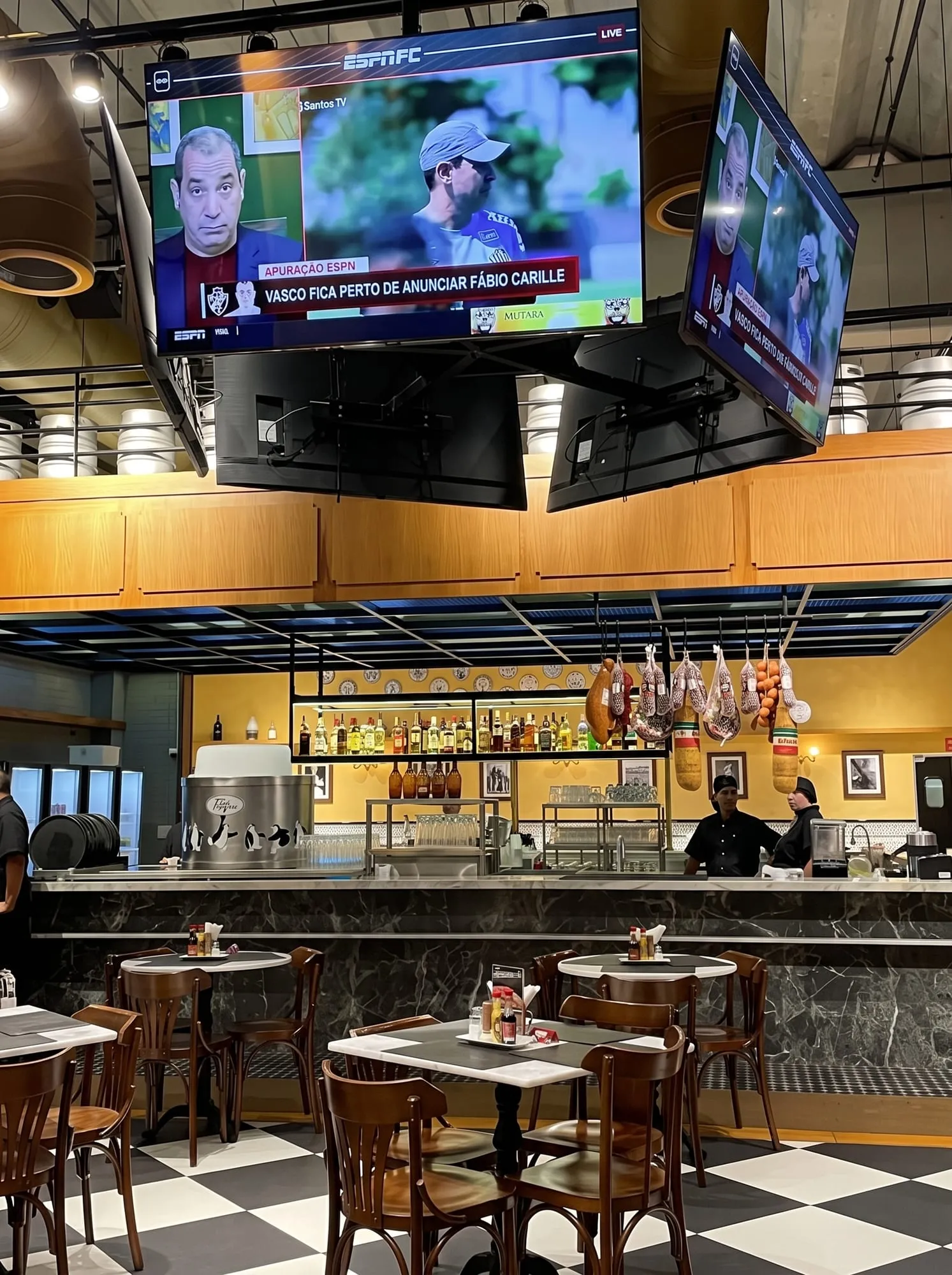 Salão da Choperia Pinguim com o bar ao fundo, TVs e piso xadrez